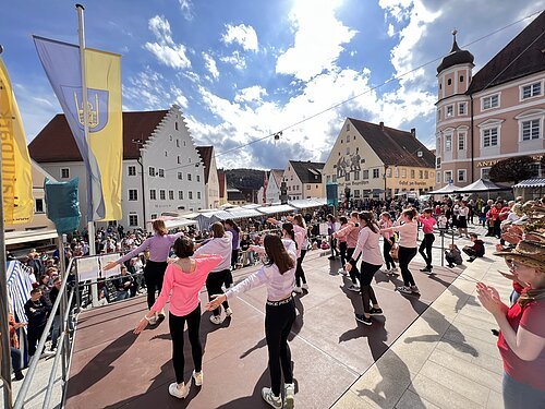 Tanzende Personen tanzen auf einer Bühne. Im Hintergrund die Altstadtkulisse von Greding, die den Rahmen des Marktplatzes bildet auf dem Marktstände und viele Besucher zu sehen sind.