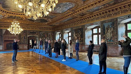 Personengruppe in einem prunkvollen Raum mit verzierten Wänden und Decken, zwei Kronleuchtern und blauen Teppichen.