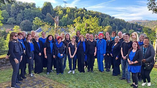 NewWay Voices Gruppenfoto von 30 Personen im Freien vor bewaldeten Hügeln bei Tageslicht.