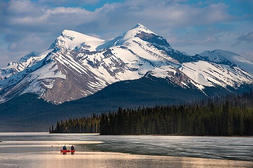 Zwei Personen in einem roten Kanu auf einem See vor schneebedeckten Bergen und einem Nadelwald.
