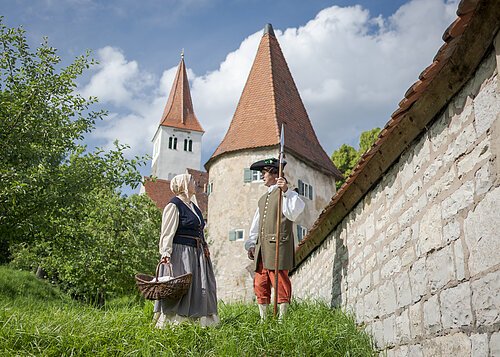 Frau mit Korb und Mann mit Hellebarde in historischer Kleidung vor Turm und Mauer bei blauem Himmel