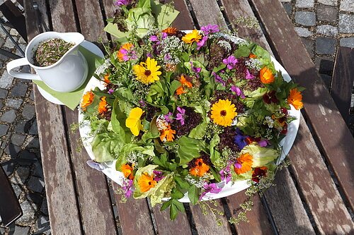 Kräuterblumensalat Bunter Salat mit essbaren Blüten auf weißem Teller neben weißem Krug mit Kräuterdressing auf Holztisch.